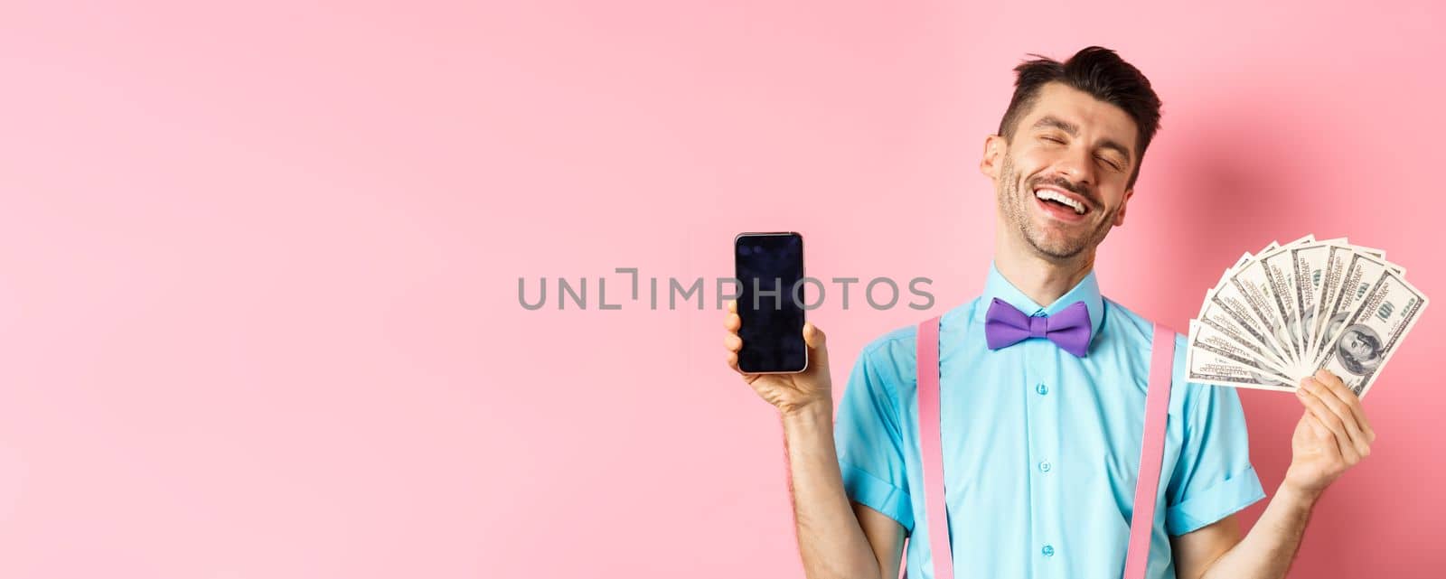 E-commerce and shopping concept. Rich and happy guy laughing, showing money in cash and empty smartphone screen, standing on pink background.