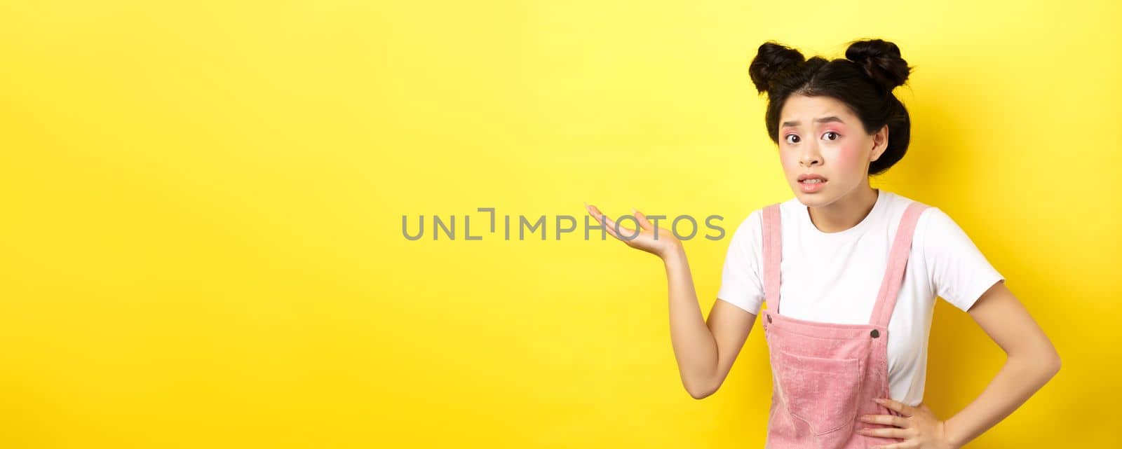 Confused asian teen girl with bright makeup, raising hand up clueless, looking puzzled, cant understand something, pointing at copy space, yellow background by Benzoix