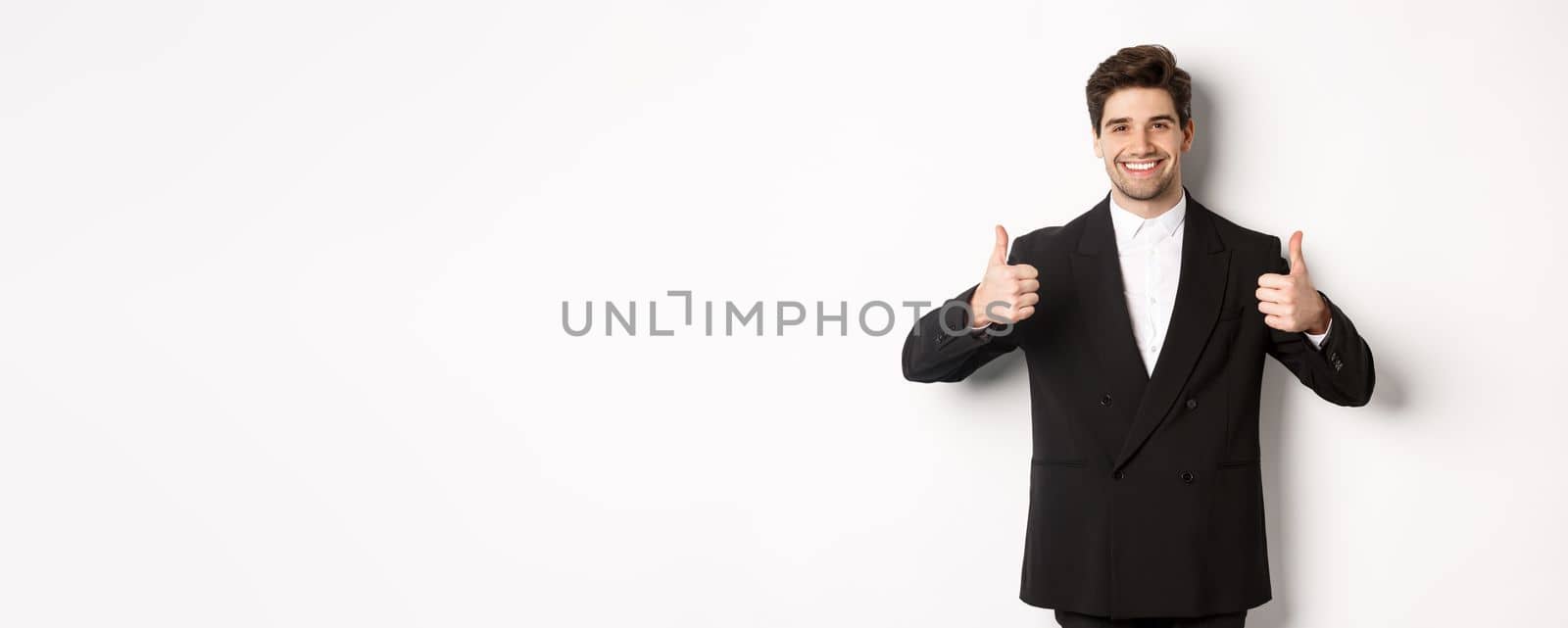 Concept of new year party, celebration and lifestyle. Image of attractive happy businessman in formal suit, showing thumbs-up and smiling, like and approve, standing over white background by Benzoix