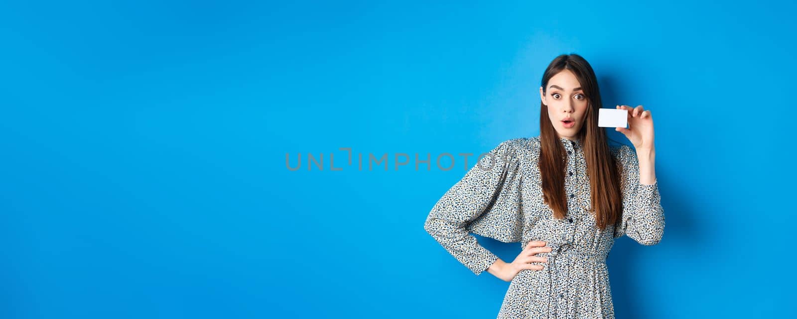 Shopping. Excited girl showing her plastic credit card and look at discounts, say wow amazed, standing on blue background.