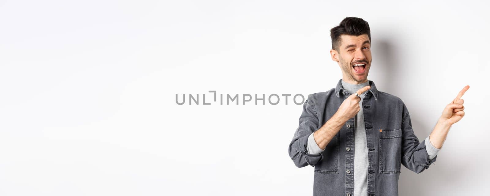 Handsome smiling guy making announcement, winking and pointing at upper right corner, showing logo, inviting to check out advertisement, standing on white background by Benzoix