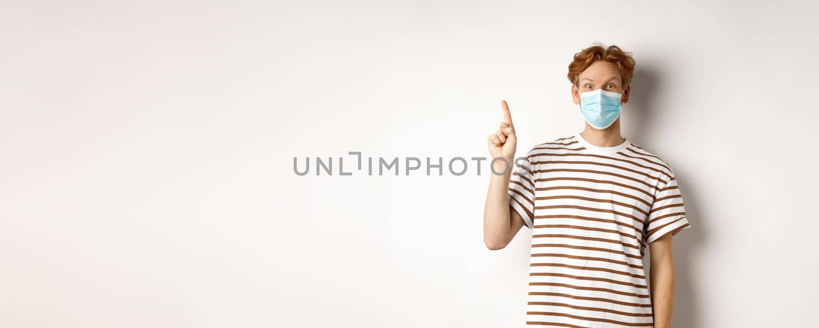 Covid, virus and social distancing concept. Surprised redhead man in face mask showing advertisement on top, pointing finger up and look amazed, white background by Benzoix