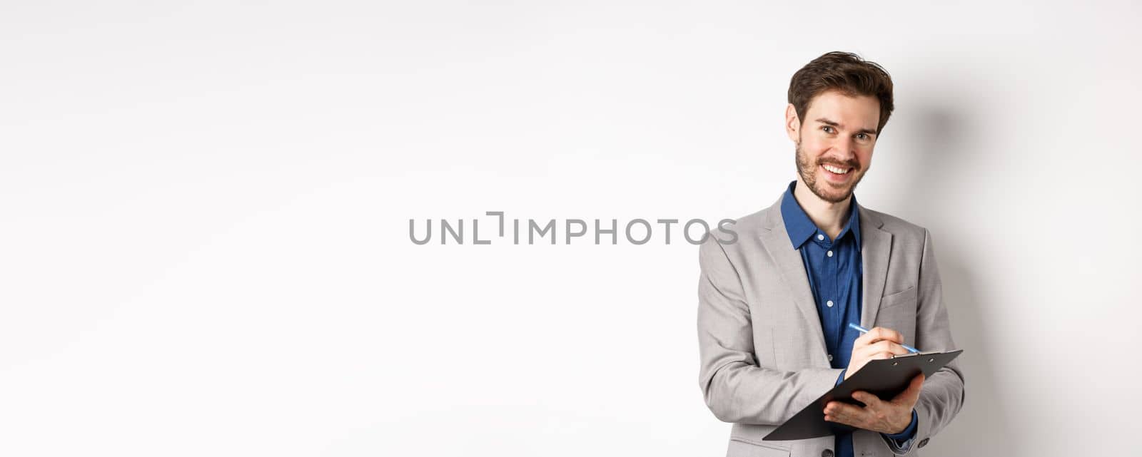 Smiling male manager in suit writing on clipboard, taking notes at office meeting, standing on white background by Benzoix