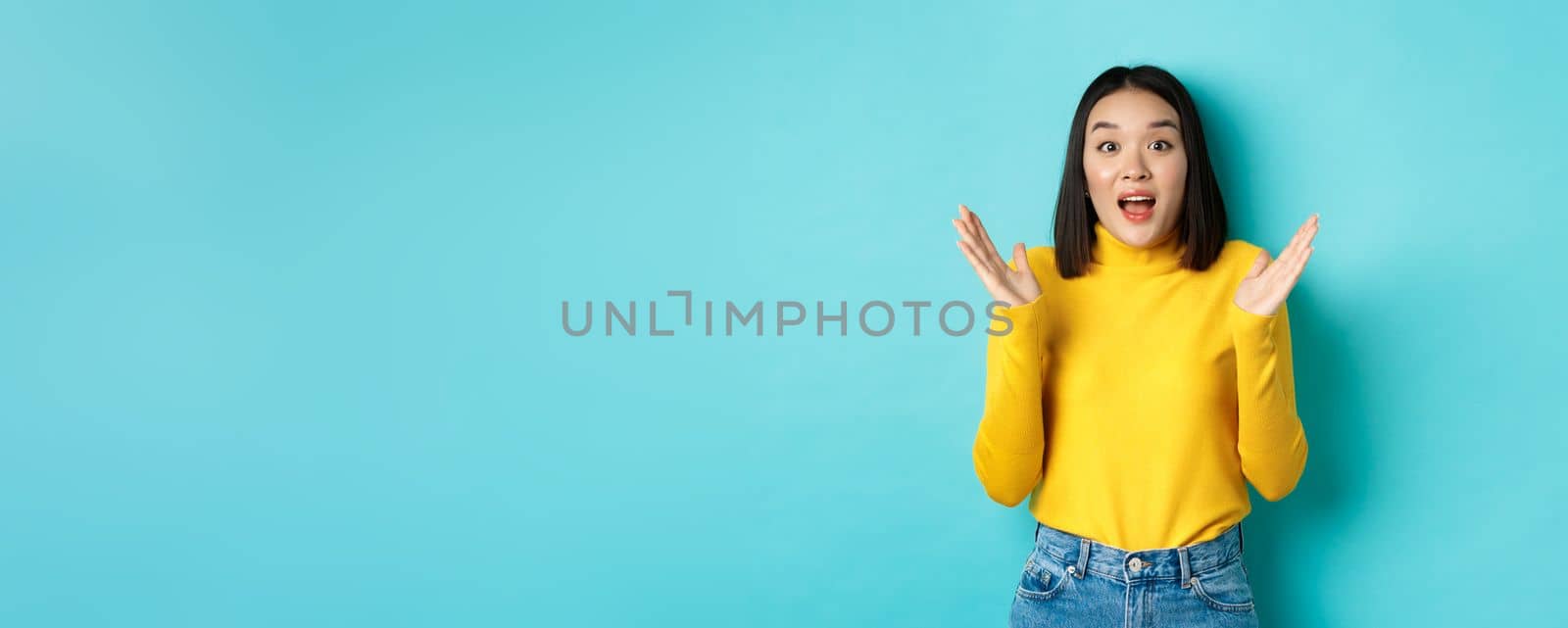 Image of happy asian woman clap hands and looking amazed, standing in yellow pullover over blue background.