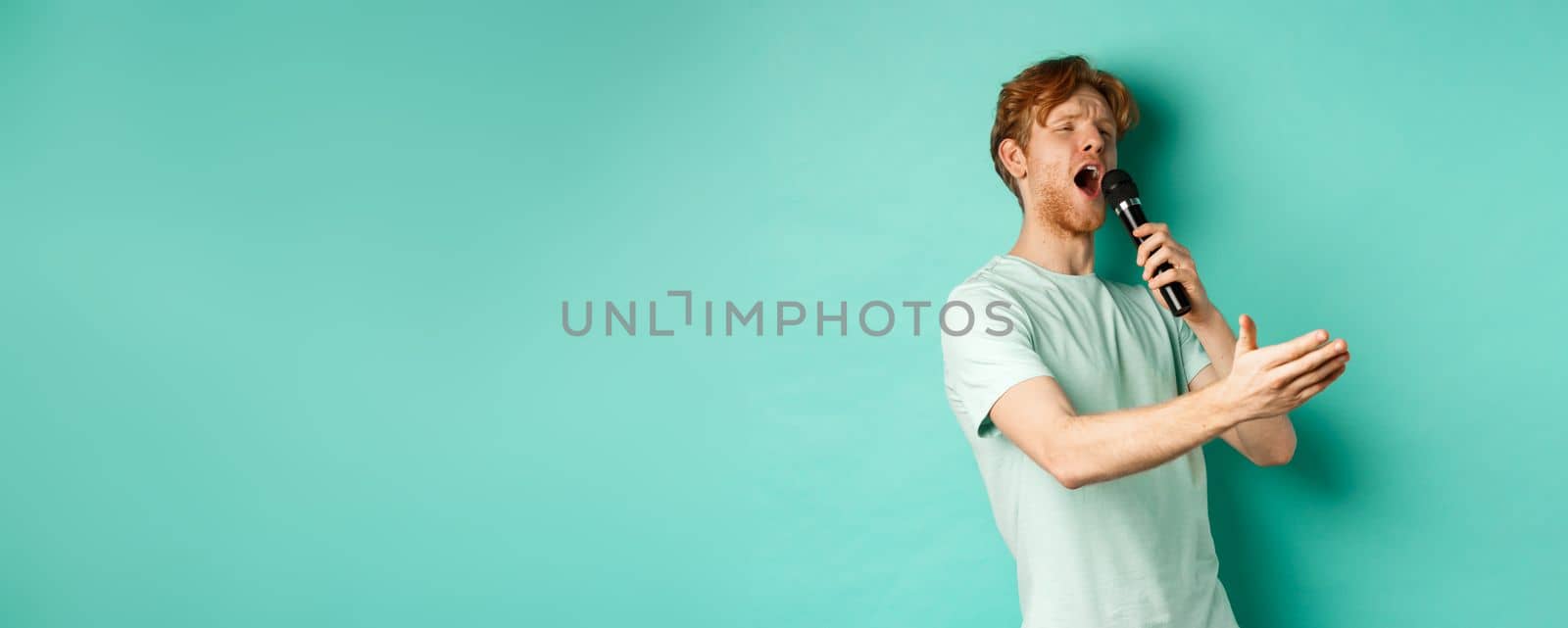 Passionate redhead man in t-shirt singing serenade with microphone, looking aside at karaoke and gesturing, standing over mint background by Benzoix