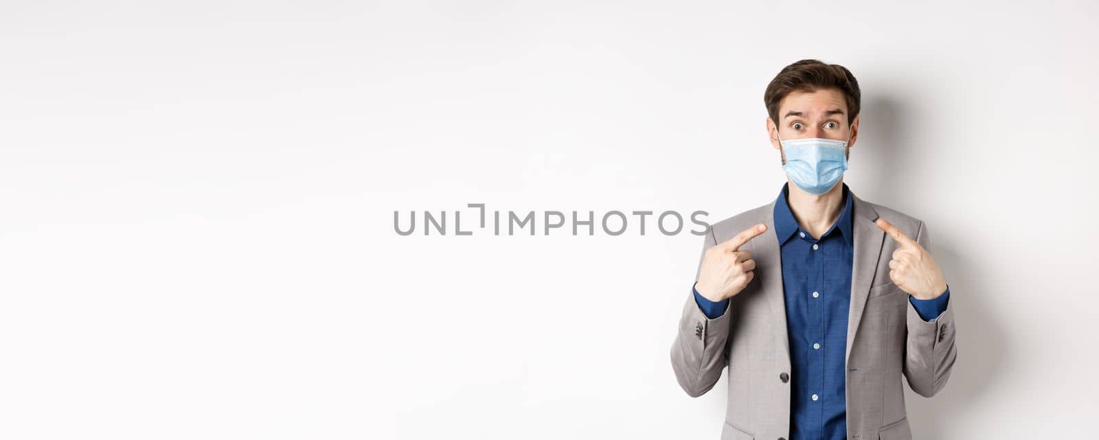 Covid-19, pandemic and business concept. Excited businessman in suit pointing at his medical mask and looking at camera, white background by Benzoix