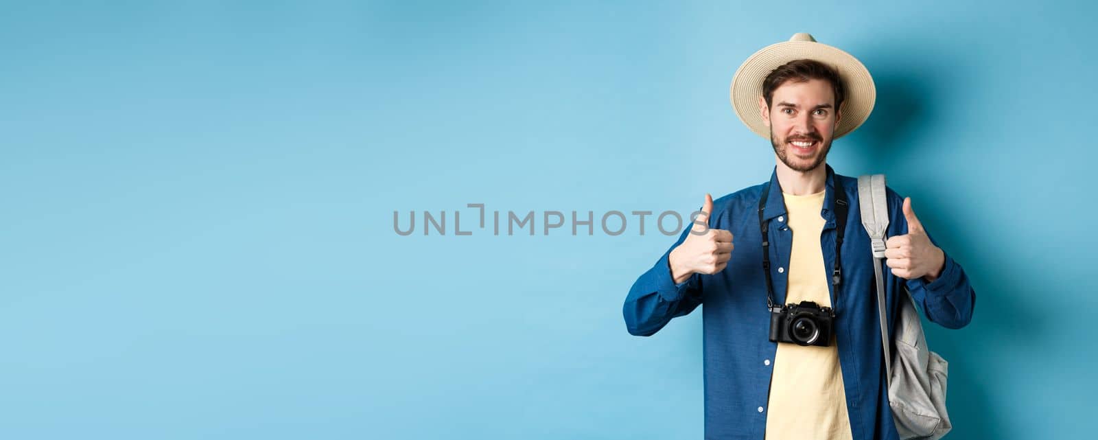 Cheerful handsome man recommending summer vacation place, showing thumbs up and smiling in approval. Tourist leave positive feedback, standing with camera and backpack on blue background.