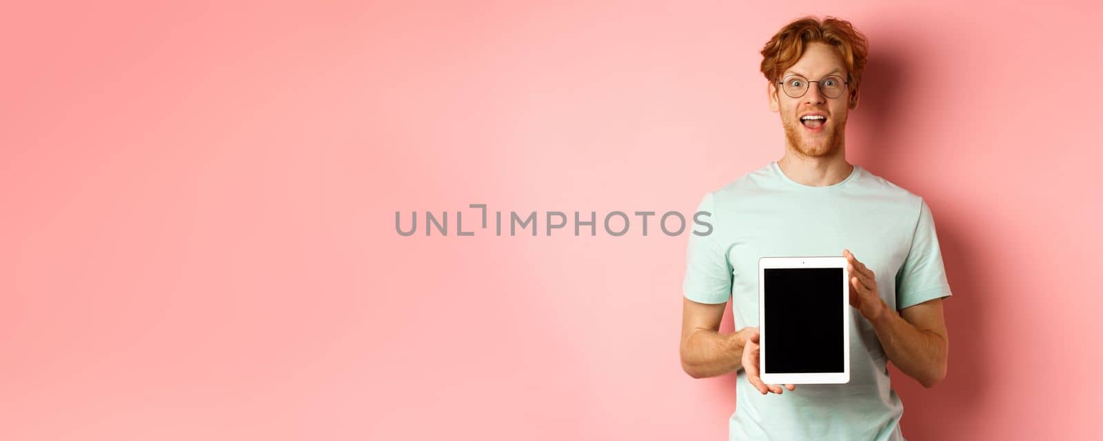 Excited young man with red hair and beard, checking out online promotion, showing digital tablet screen and staring at camera amazed, standing over pink background.