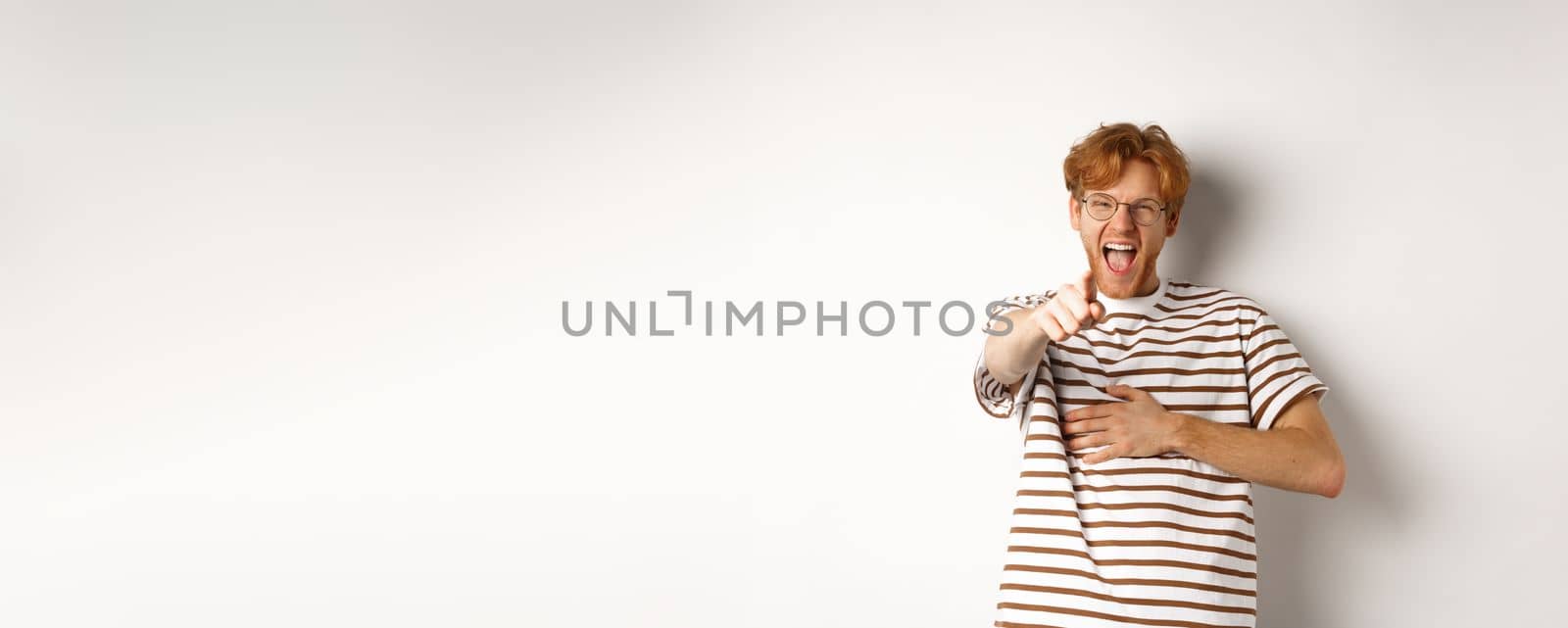Young man with ginger hair and beard pointing finger at camera and laughing, making fun of someone hilarious, standing over white background by Benzoix