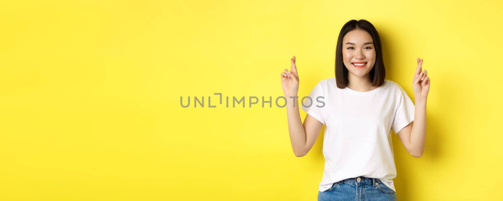 Hopeful asian girl making wish, cross fingers for good luck and praying with eyes closed, standing over yellow background.