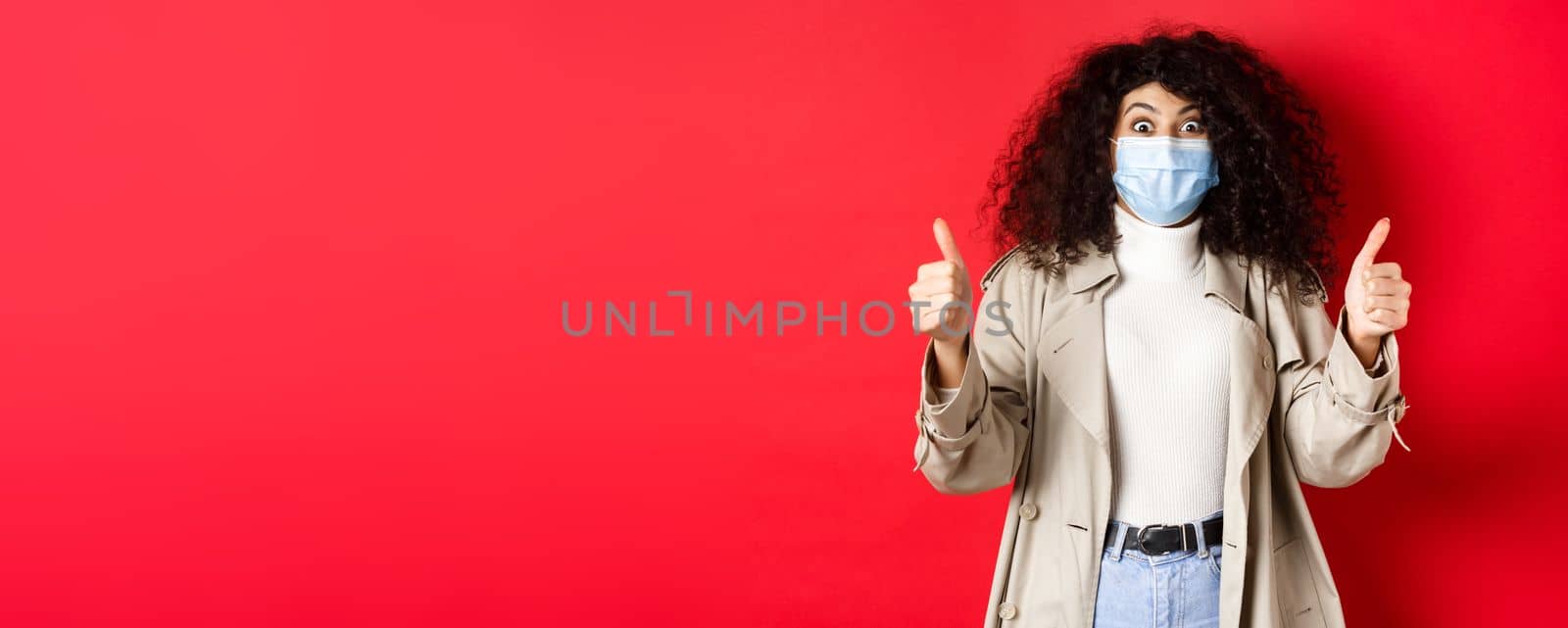 Covid-19, pandemic and quarantine concept. Excited girl with curly hair, wearing trench coat and medical mask, showing thumbs up in approval, red background.