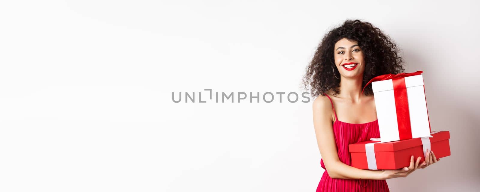 Beautiful birthday girl with curly hair, holding bday gifts and smiling happy, celebrating, standing against white background.