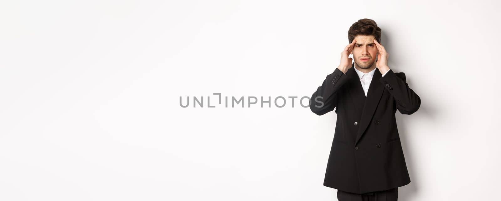 Image of businessman in black suit, touching head and looking dizzy, feeling painful headache, standing over white background.