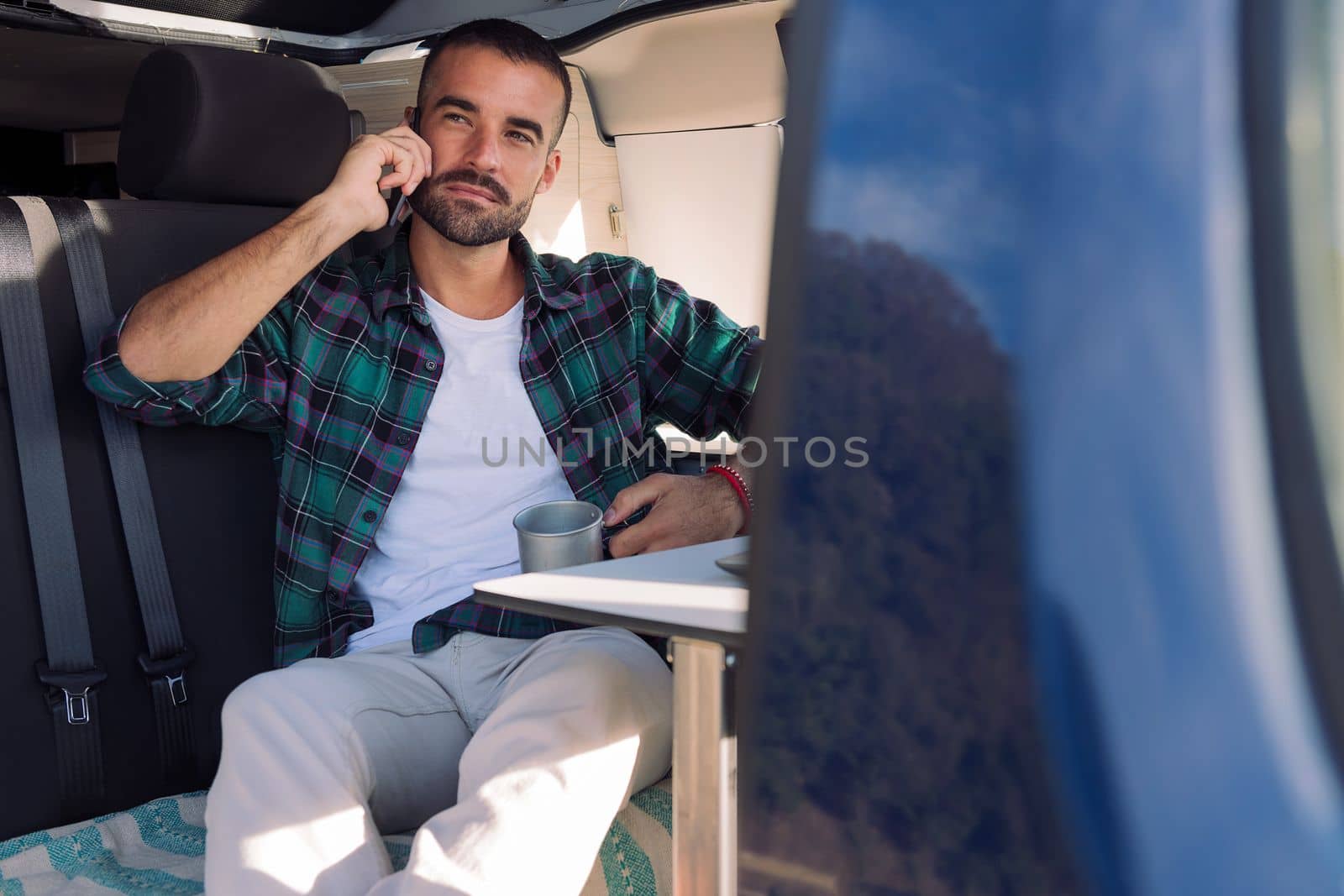 young man sitting inside his camper van talking on mobile phone, concept of freedom and digital nomad lifestyle