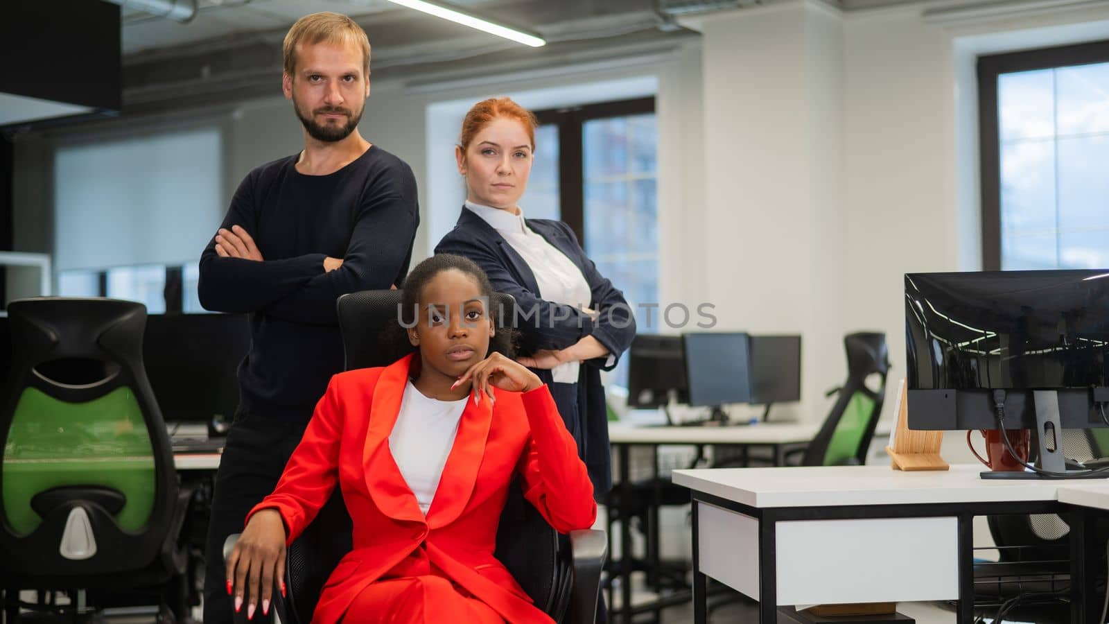 Caucasian red-haired woman, bearded Caucasian man stand behind a seated African American young woman in the office. by mrwed54