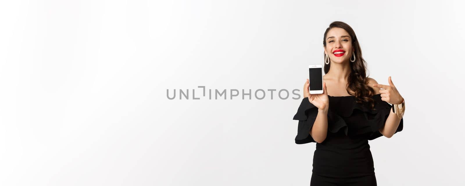 Online shopping concept. Fashionable woman in black dress pointing finger at smartphone screen, showing application, standing over white background by Benzoix