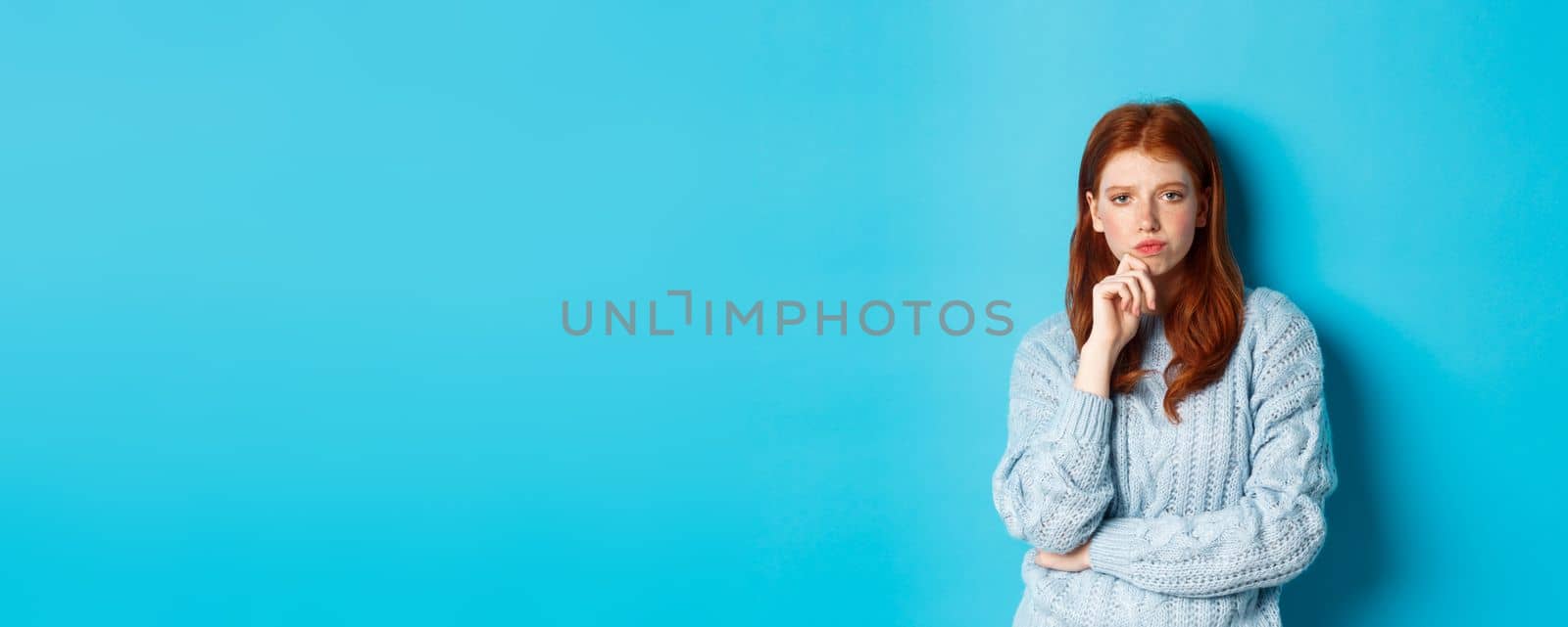 Thoughtful teenage redhead girl staring at camera, pondering ideas, making decision with serious face, standing over blue background.