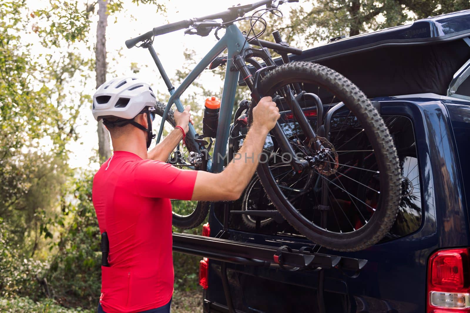 rear view of a young man in helmet and cycling clothes taking his mountain bike off the bike rack of his van to go for a training ride, concept of freedom and sport in nature