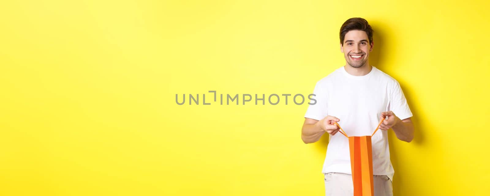 Happy young man open shopping bag with present, smiling at camera, standing against yellow background.