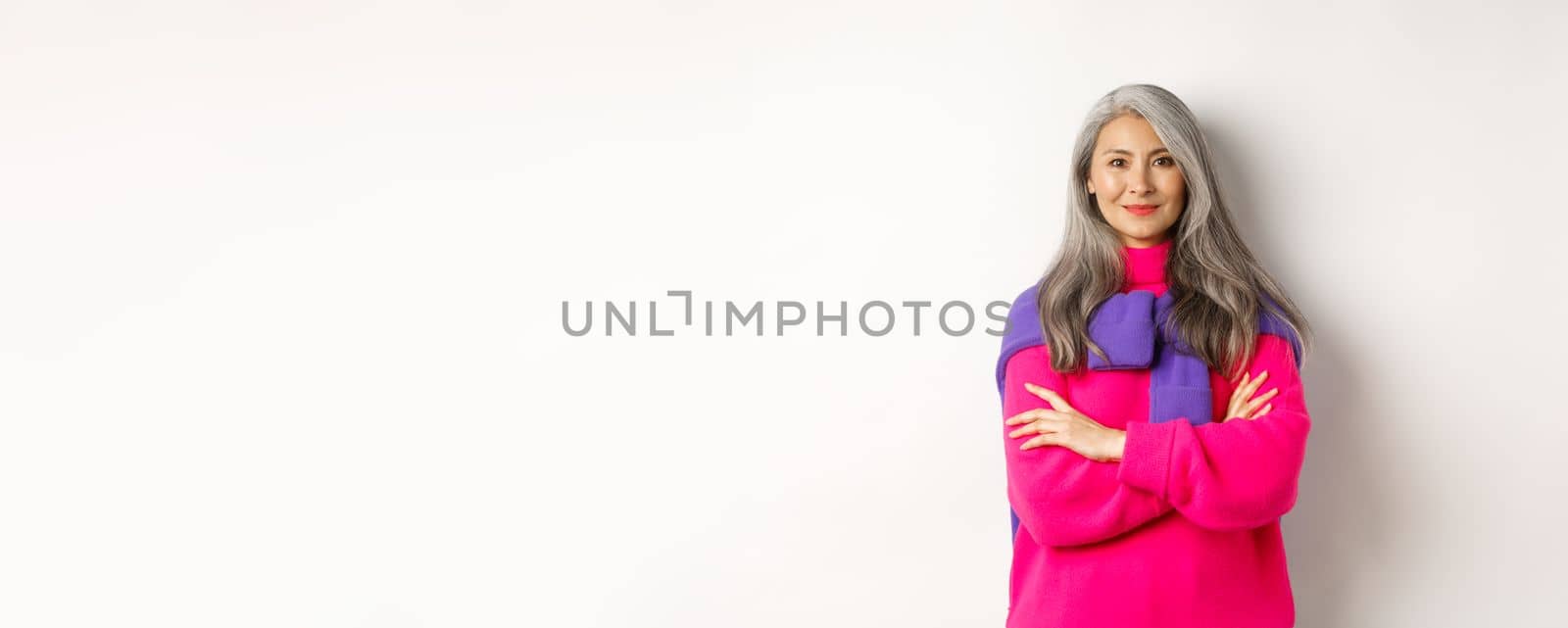 Portrait of confident asian senior woman cross arms on chest and smiling, standing over white background in trendy clothes by Benzoix