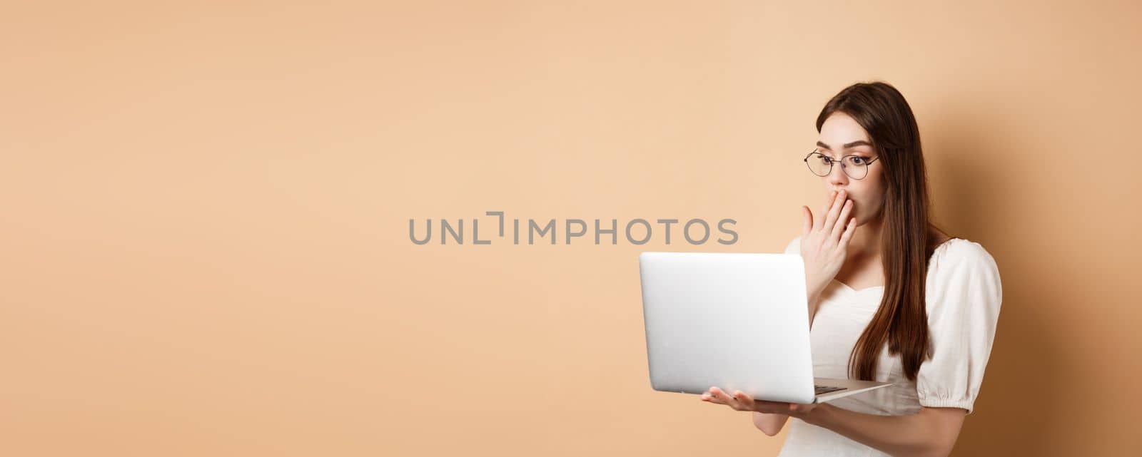 Shocked girl look at laptop screen, covering mouth with amazed face, standing in glasses on beige background.