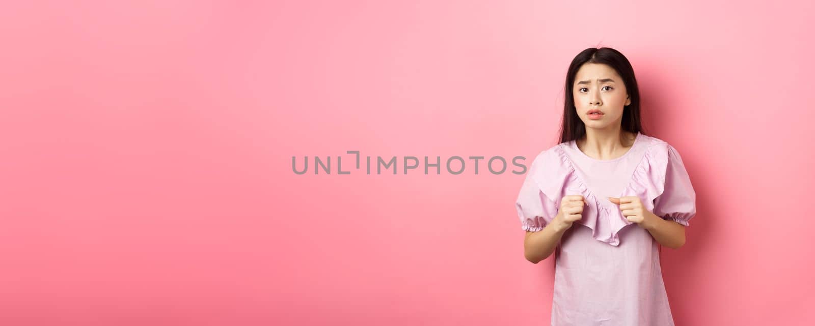 Scared teen asian girl clench fists and looking alarmed at camera, frightened with something scary, standing on pink background.