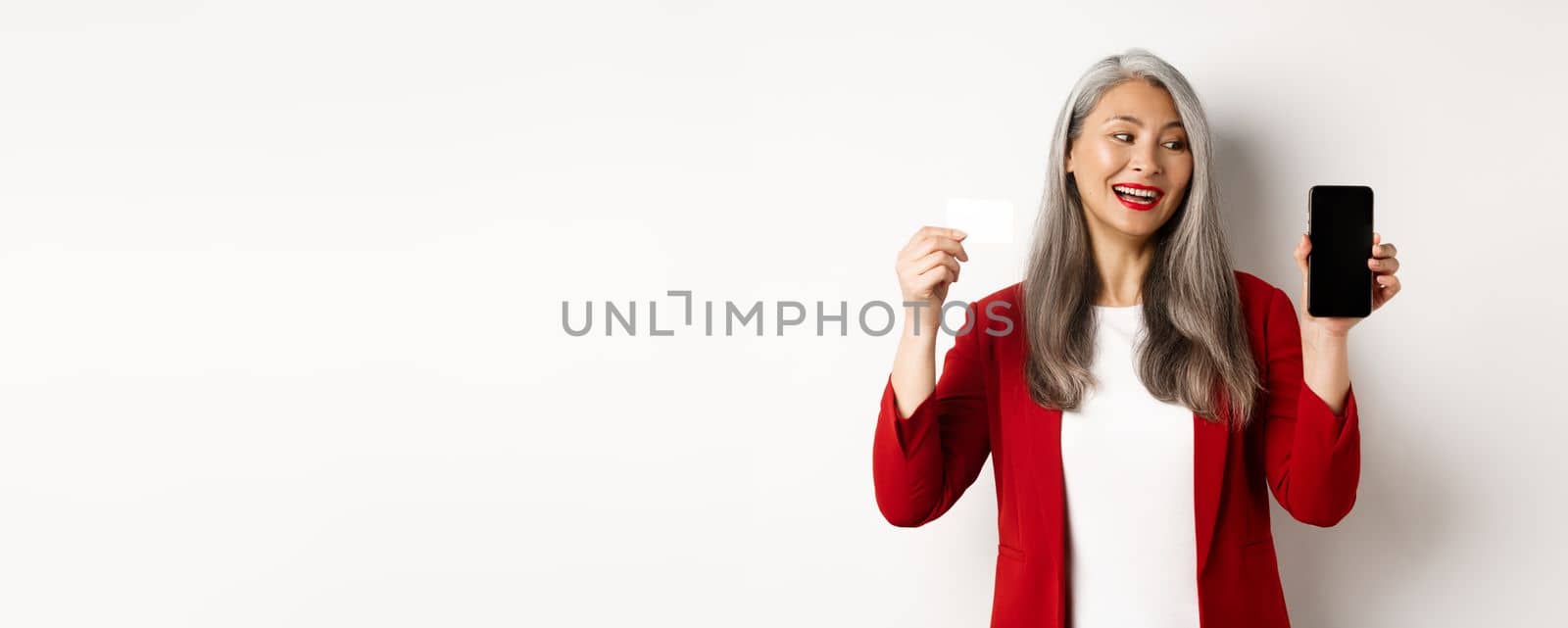 Cheerful asian mature woman showing blank smartphone screen and credit card, concept of e-commerce.