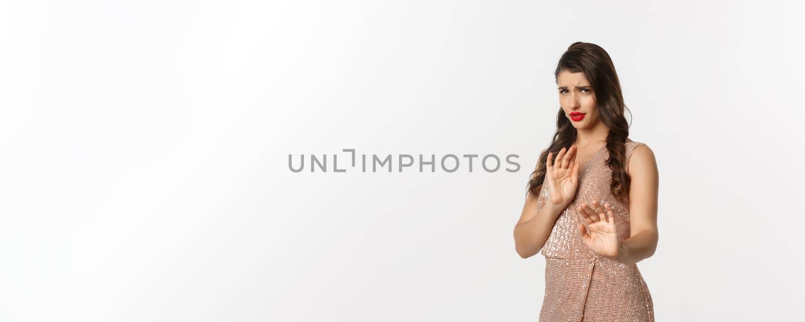 Concept of celebration, holidays and party. Woman looking displeased and rejecting something, wearing glamour dress, showing stop gesture, asking stay away, white background.