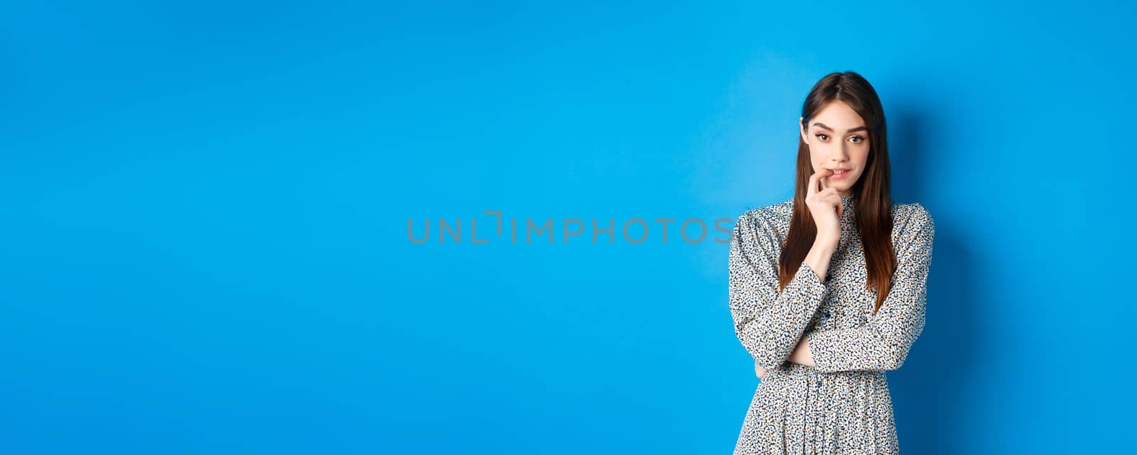 Sensual woman in dress biting finger and looking pensive at camera, standing dreamy against blue background.
