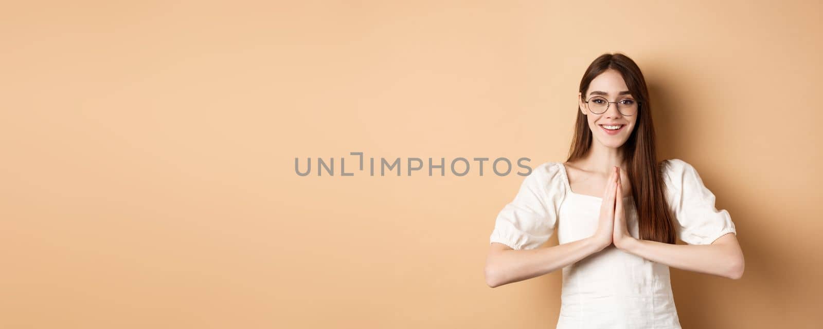 Smiling girl in glasses say thank you, show namaste gesture, express gratitude, standing pleased on beige background.