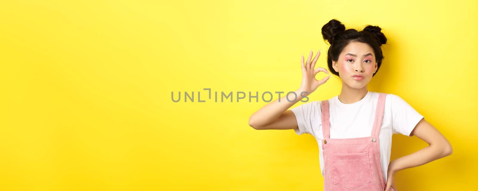 Sassy teenage asian girl with bright pink makeup, showing okay sign and nod in approval, approve product with serious face, not bad gesture, standing against yellow background by Benzoix
