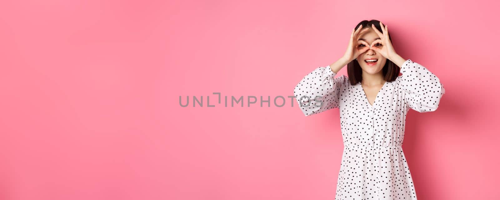 Carefree young asian woman looking through hand binoculars at camera, staring at discounts, standing over pink background.