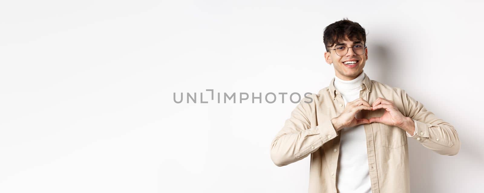 Happy Valentines. Handsome young man in glasses say I love you, showing heart sign and smiling at girlfriend, celebrating lovers day, standing over white background.