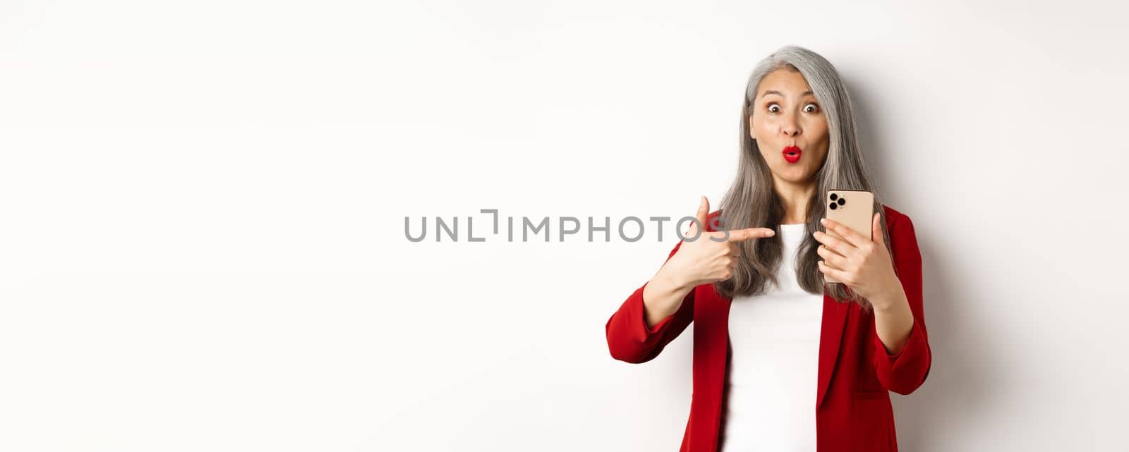 Amazed korean lady in red blazer pointing finger at mobile phone, looking curious at camera, white background.