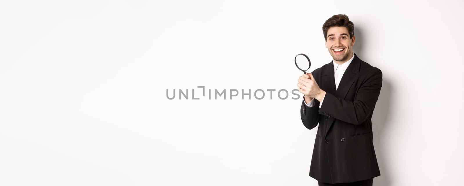 Handsome businessman in black suit, holding magnifying glass and smiling, looking for you, standing against white background.