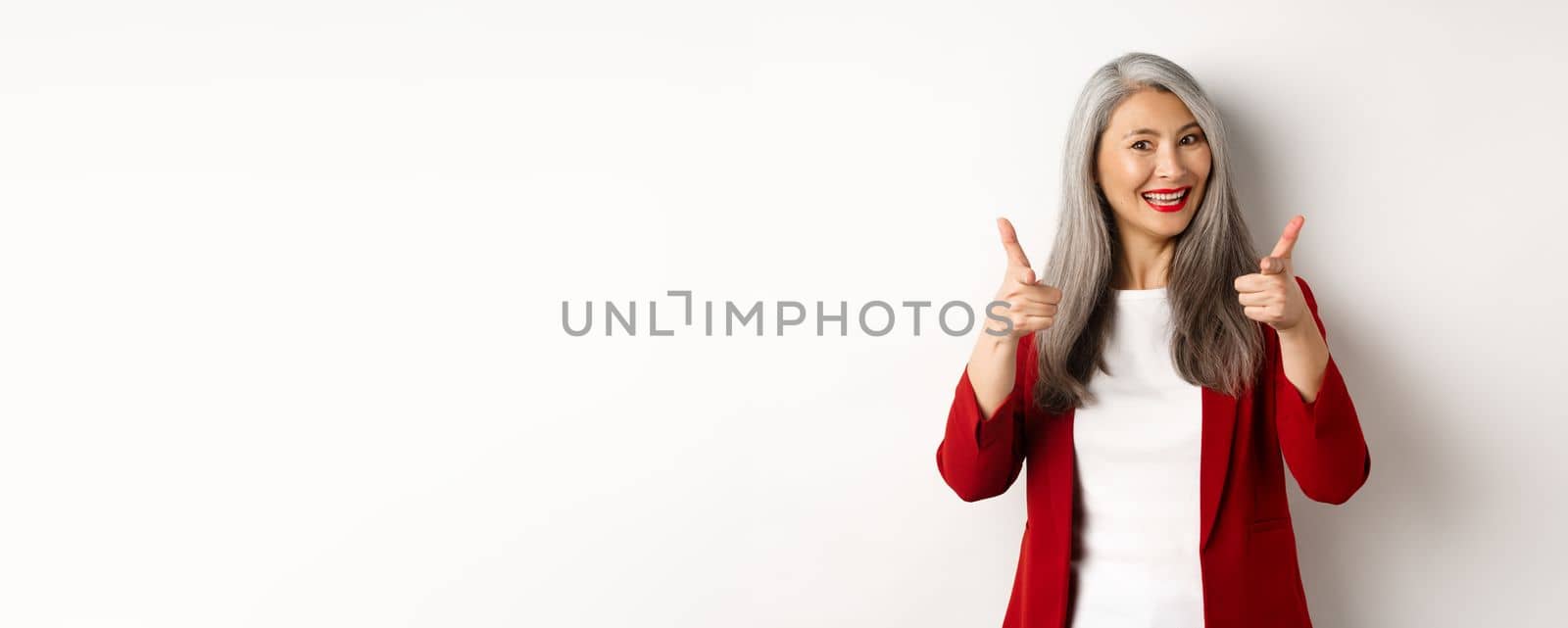 Professional female employer in trendy red blazer and makeup, pointing fingers at camera and smiling, praising something, need you, standing over white background.