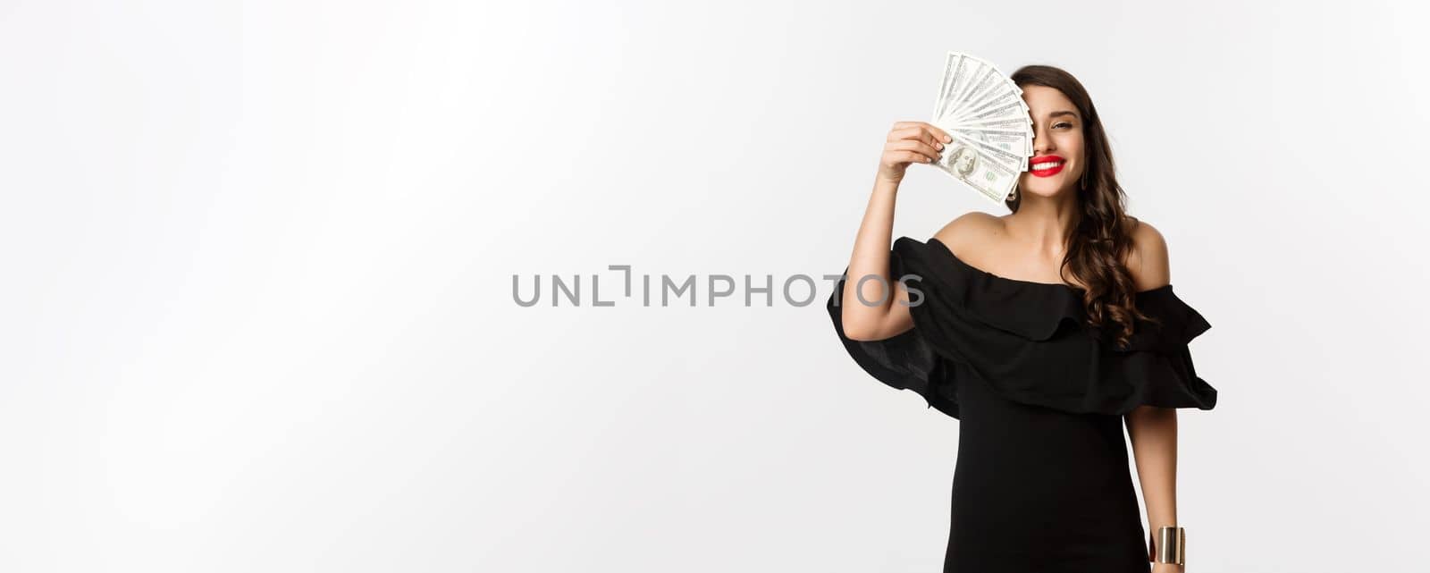 Fashion and shopping concept. Happy young woman in black dress, with red lips, holding money and smiling satisfied, standing over white background.