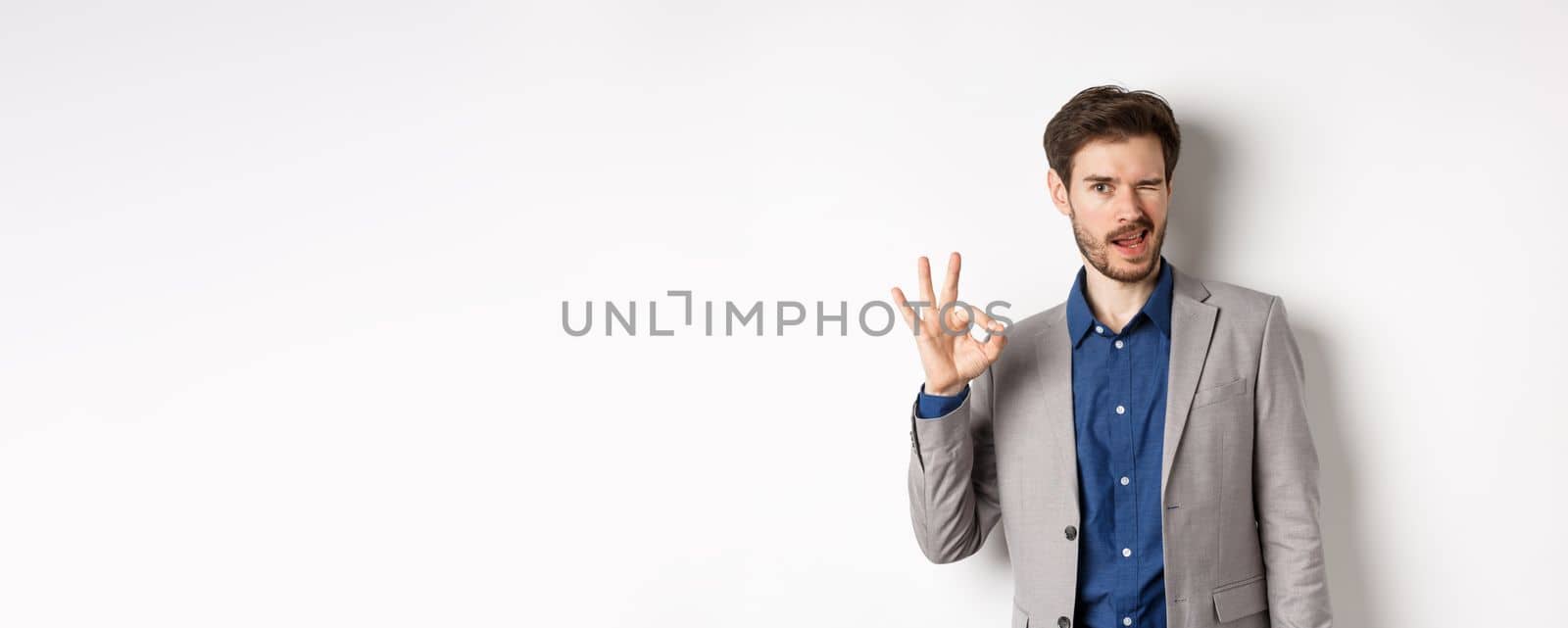 All good. Confident male ceo manager showing okay sign and winking, everything under control, praising nice work, standing on white background.
