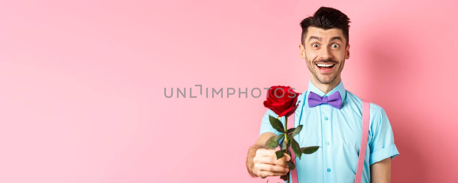 Valentines day and romance concept. Happy smiling man giving you red rose on romantic date, standing on pink background in bow-tie and shirt.