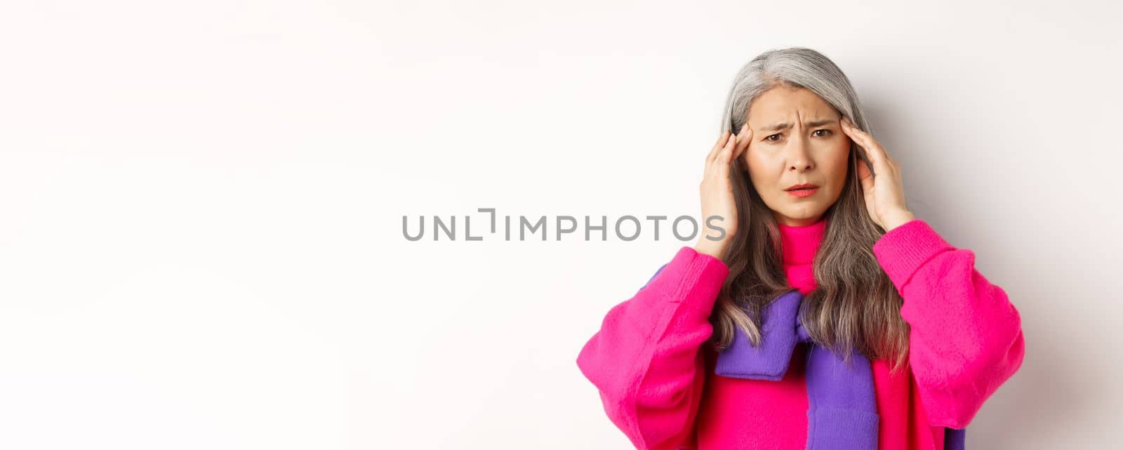Close-up of fashionable asian senior woman feeling unwell, touching head and grimacing from headache, complain on migraine, white background by Benzoix