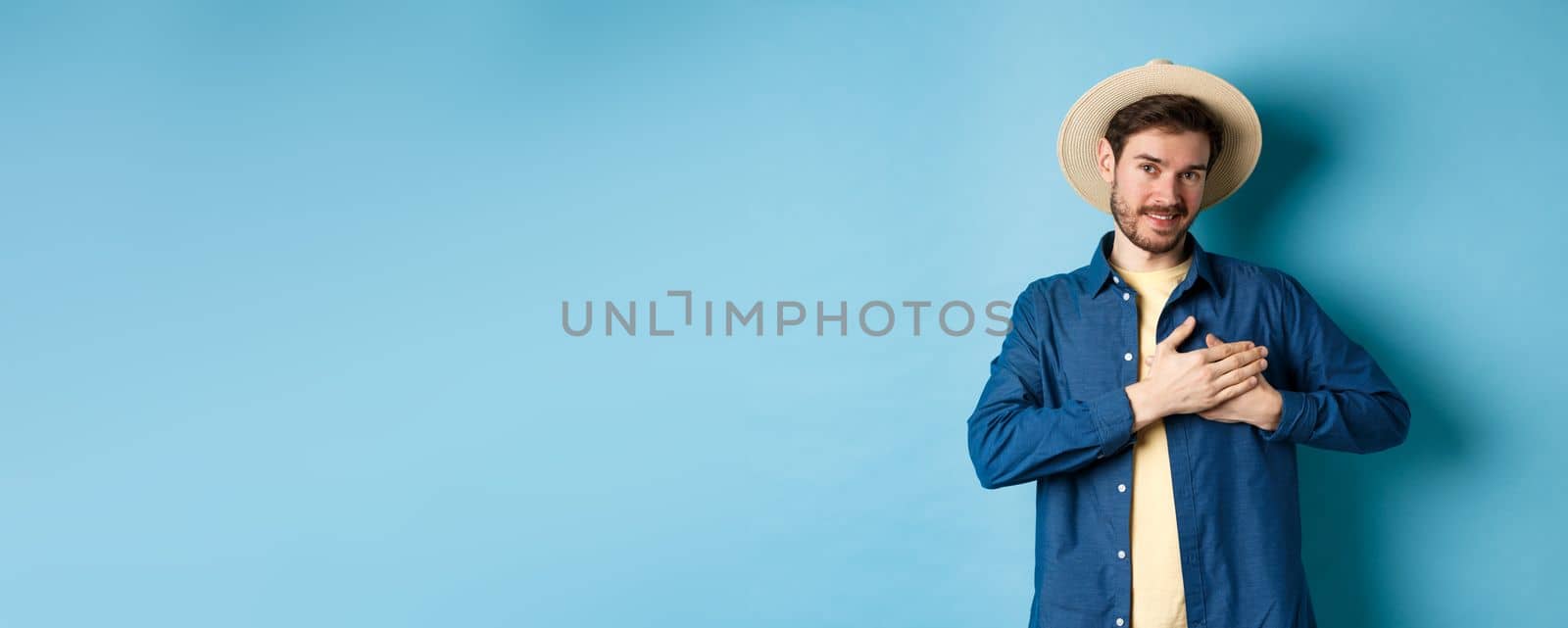 Handsome traveler in straw hat, holding hands on heart and smiling, showing thank you gesture, keep memories about vacation in soul, standing on blue background.