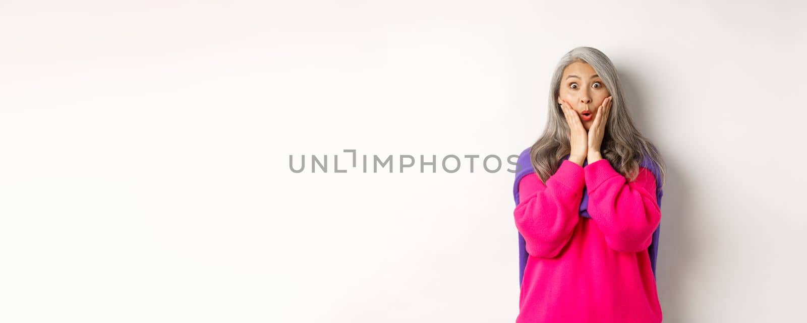 Surprised asian senior woman gasping amazed, holding hands on face and staring at camera impressed, standing over white background.