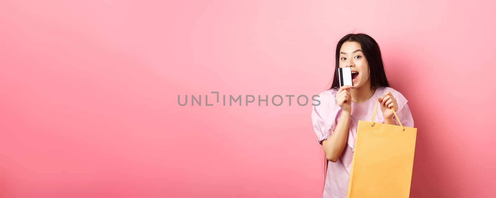 Shopping. Excited beautiful asian woman buying in stores, holding paper bag and kissing plastic credit card, standing against pink background by Benzoix