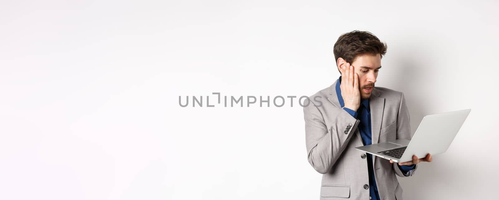Shocked and worried businessman making mistake on laptop, looking at computer upset, standing on white background.