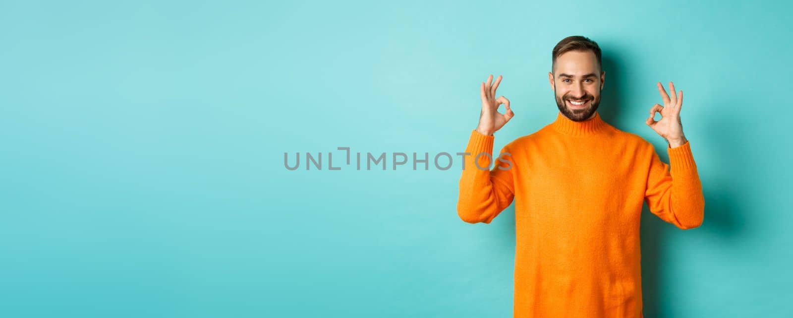 Image of confident smiling man showing okay sign, approve and agree, guarantee quality, standing over light blue background.