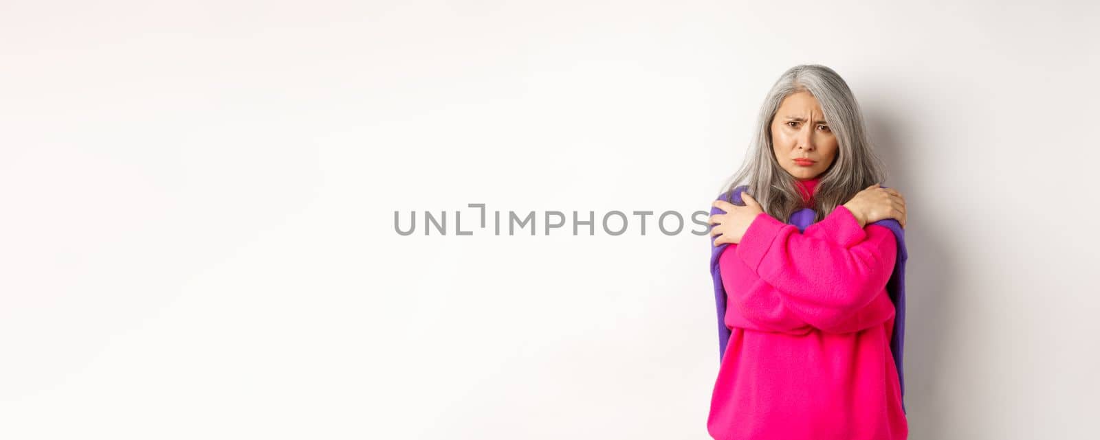 Defensive and offended asian middle-aged woman hugging body, comforting herself and looking angry at camera, frowning insulted, standing over white background.