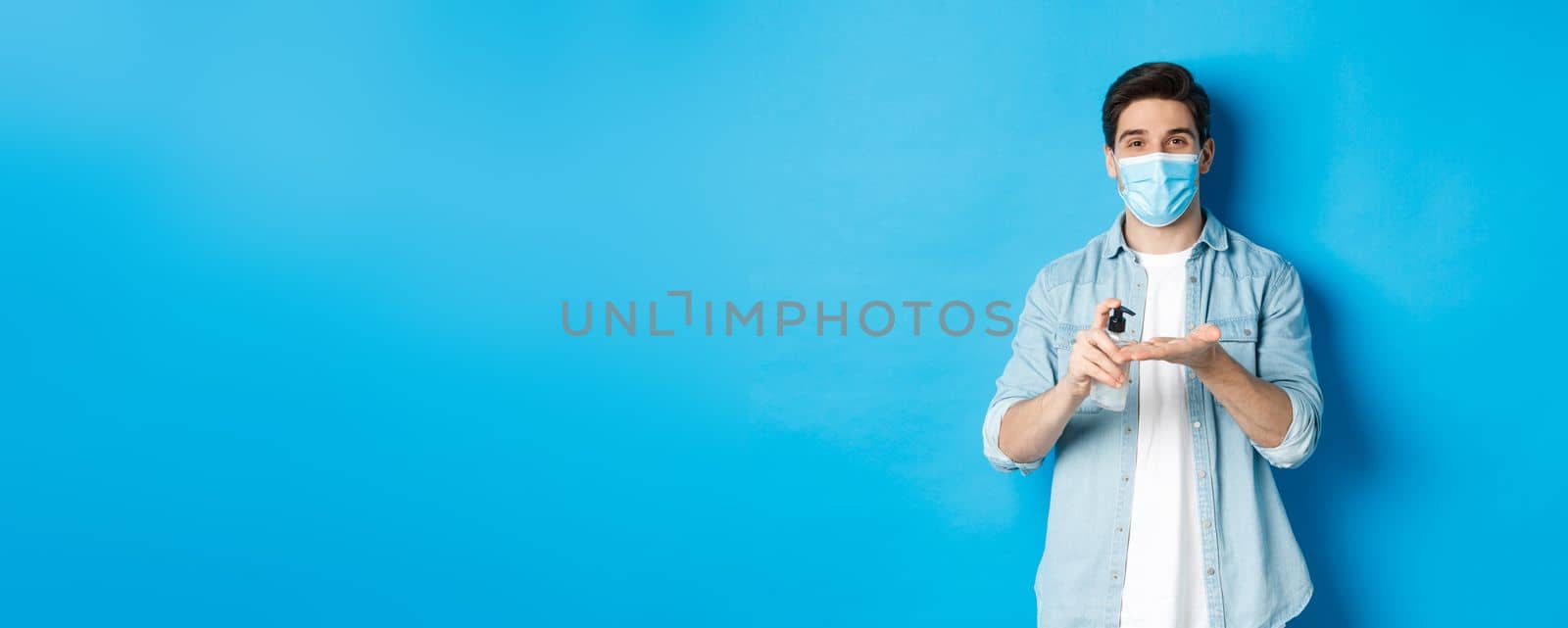 Concept of covid-19, pandemic and social distancing. Handsome young man in medical mask disinfecting hands with sanitizer, using antiseptic, standing against blue background.