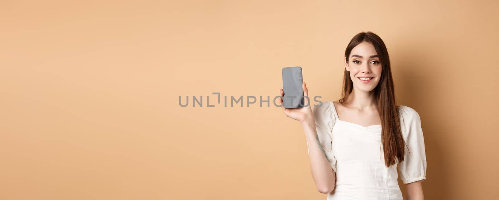 Happy girl showing empty mobile screen and smiling, demonstrate phone app, standing on beige background.