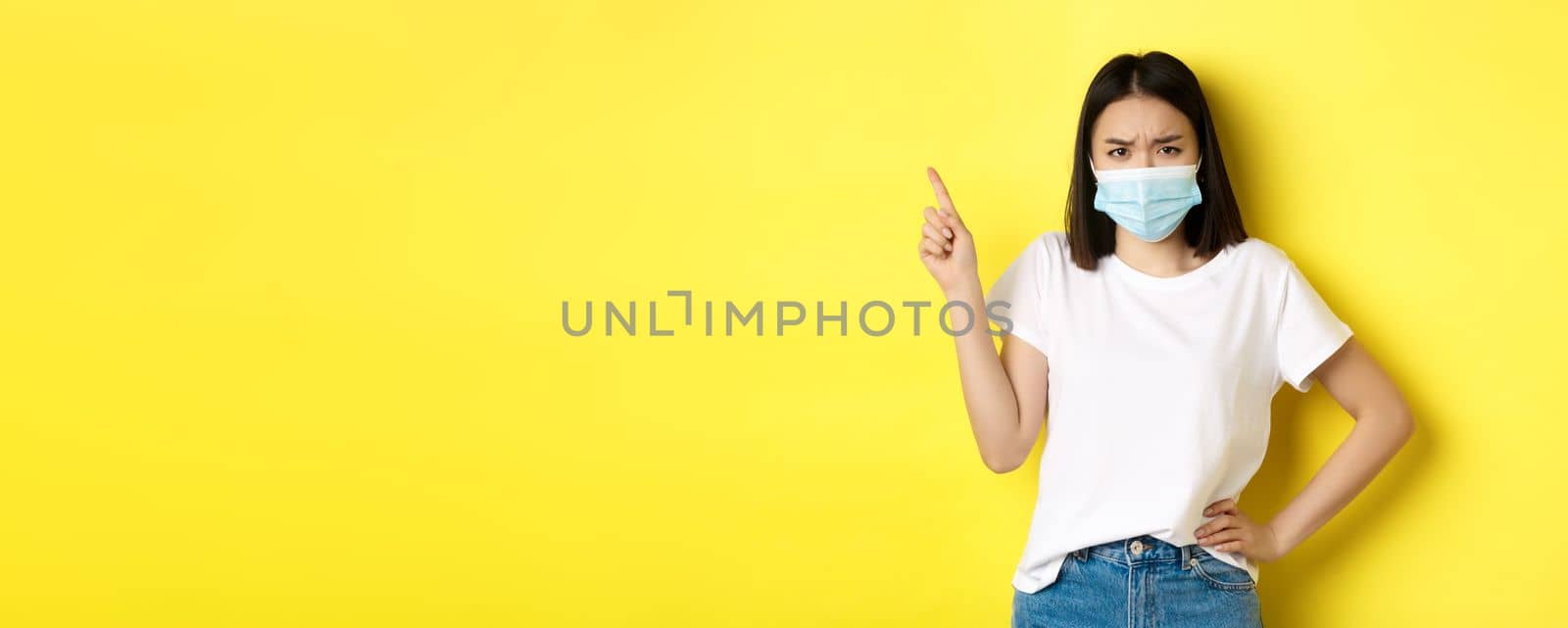 Covid, health care and pandemic concept. Asian female model in medical mask and white t-shirt pointing finger at upper left corner logo, showing promotion, yellow background.