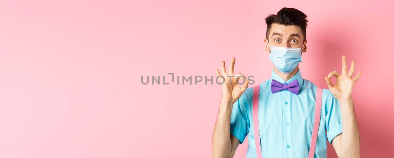 Coronavirus, healthcare and quarantine concept. Happy guy in medical mask and festive bow-tie showing all good gesture, make okay signs and smile at camera, pink background.